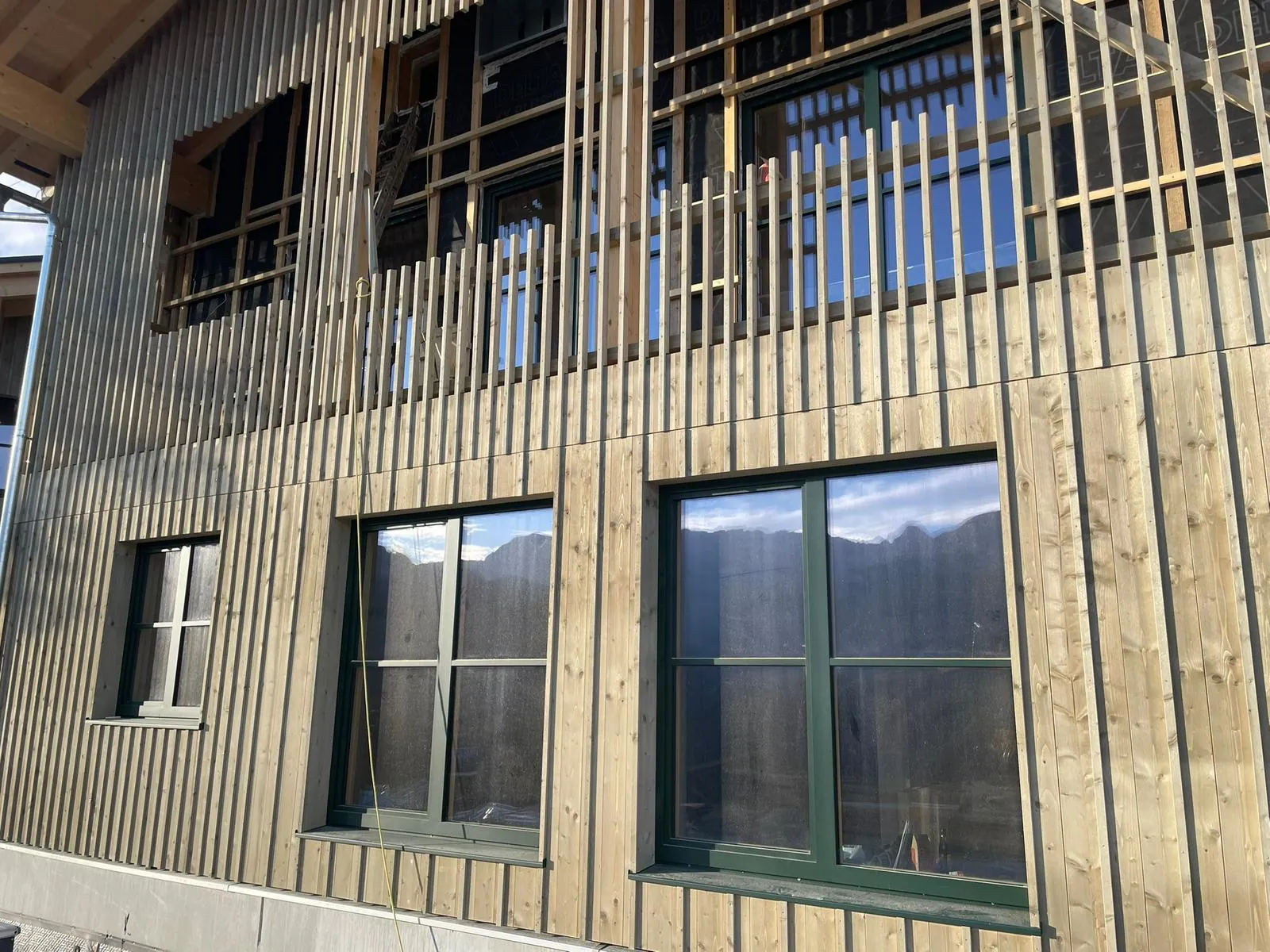 Fassadenverkleidung aus Holz mit Fenstern an einem Gebäude, im oberen Bildbereich ein Gerüst